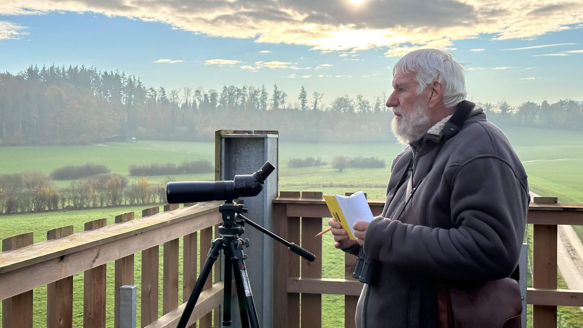 Ein älterer Mann beobachtet mit einem Fernrohr die Landschaft und hält dabei ein Notizbuch in der Hand.