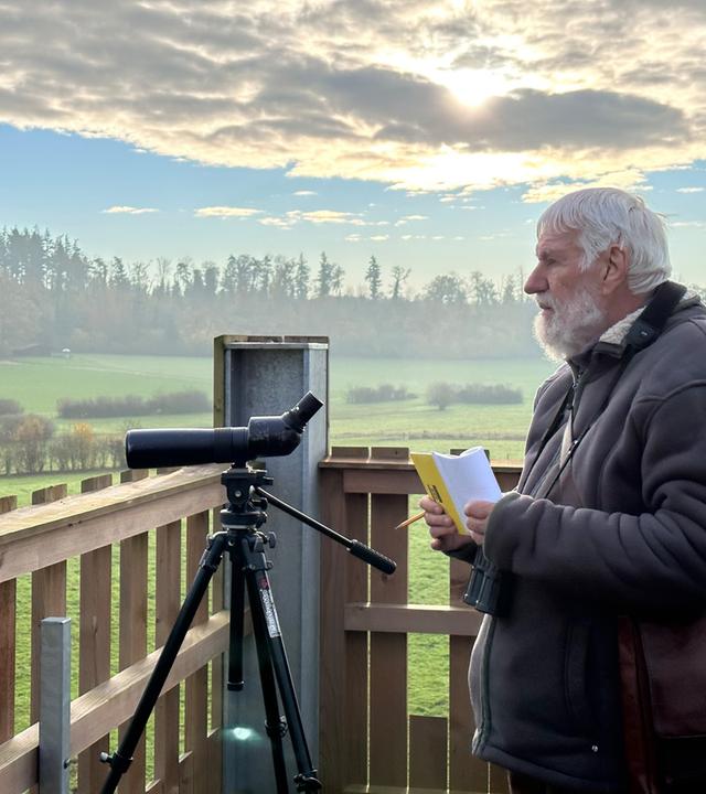 Ein älterer Mann beobachtet mit einem Fernrohr die Landschaft und hält dabei ein Notizbuch in der Hand.