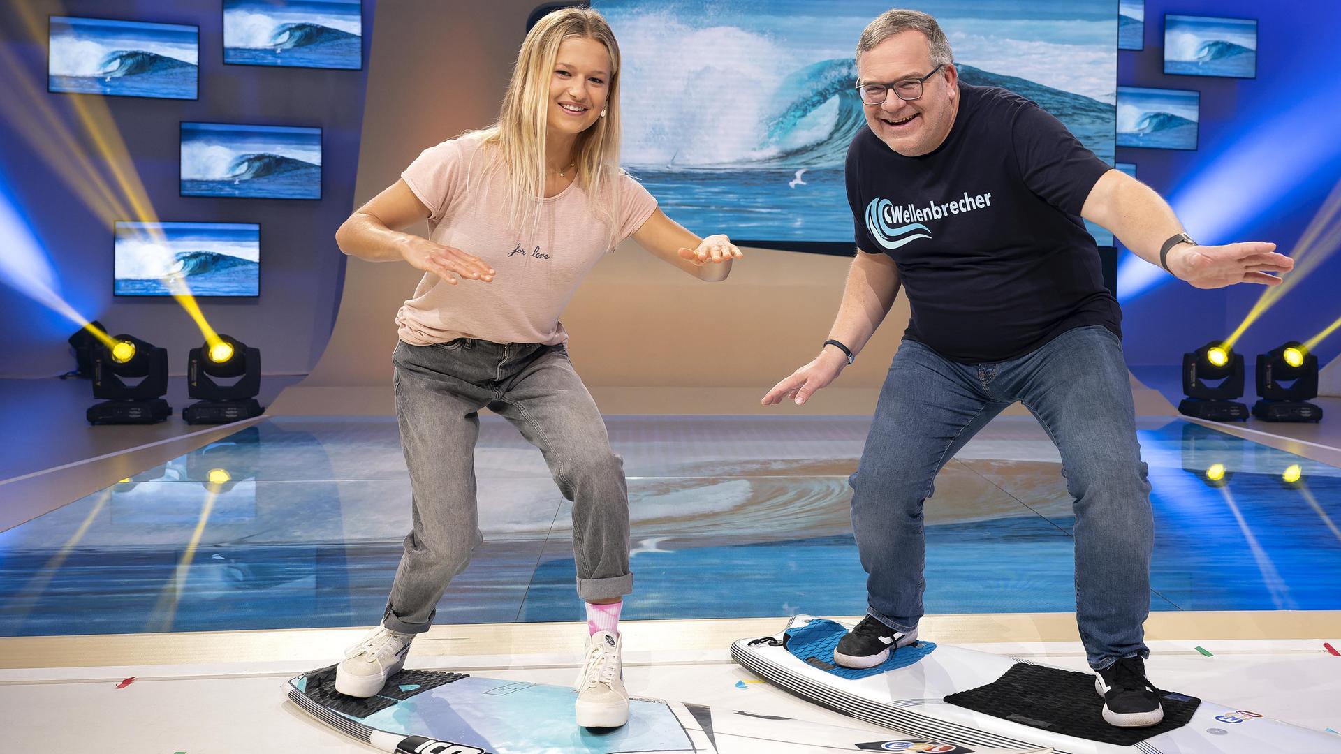 Surferin Rosina Neuerer und Elton stehen im Studio in Surfhaltung auf Surfbrettern und lachen in die Kamera.