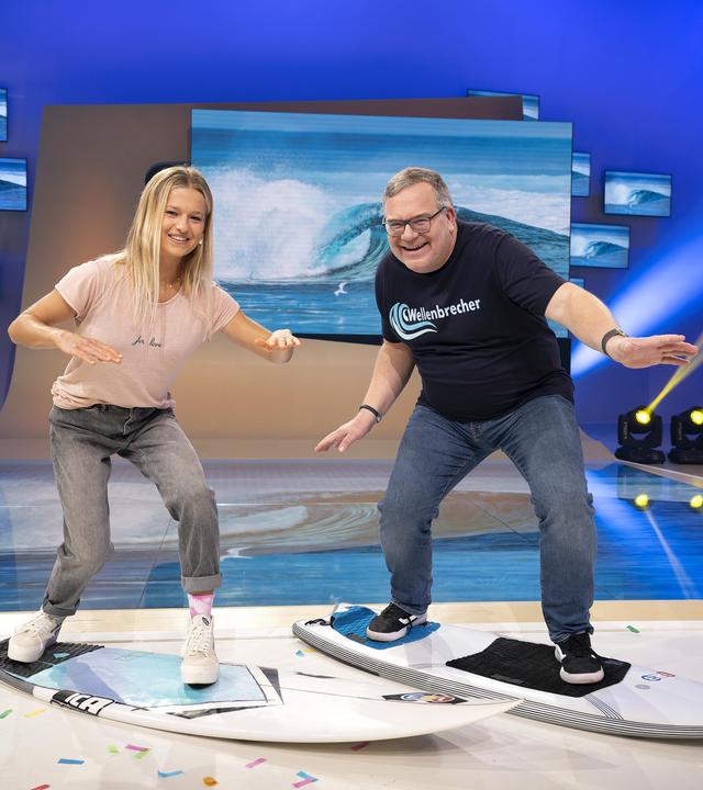Surferin Rosina Neuerer und Elton stehen im Studio in Surfhaltung auf Surfbrettern und lachen in die Kamera.