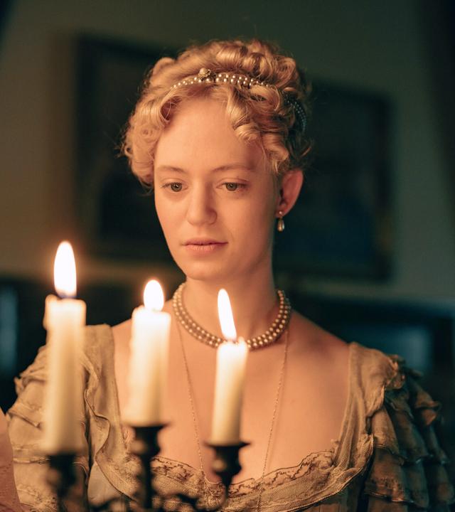 Das Bild zeigt eine Frau, die in einem historischen Raum dargestellt ist. Sie hält eine brennende Kerze in einer Hand, während sie eine andere Kerze anzündet. Die Frau trägt ein elegantes, antikes Kleid in sanften Farben mit Rüschen und einer Perlenkette um den Hals. Ihre Haare sind zu einem lockigen Chignon frisiert, und sie hat kleine Ohrringe. Der Hintergrund des Bildes ist schwach beleuchtet, was auf eine gedämpfte und intime Atmosphäre hinweist. Die brennenden Kerzen erzeugen ein warmes Licht und werfen sanfte Schatten im Raum. An den Wänden sind verschwommen Bilder oder Gemälde zu erkennen, die zur historischen Kulisse beitragen.