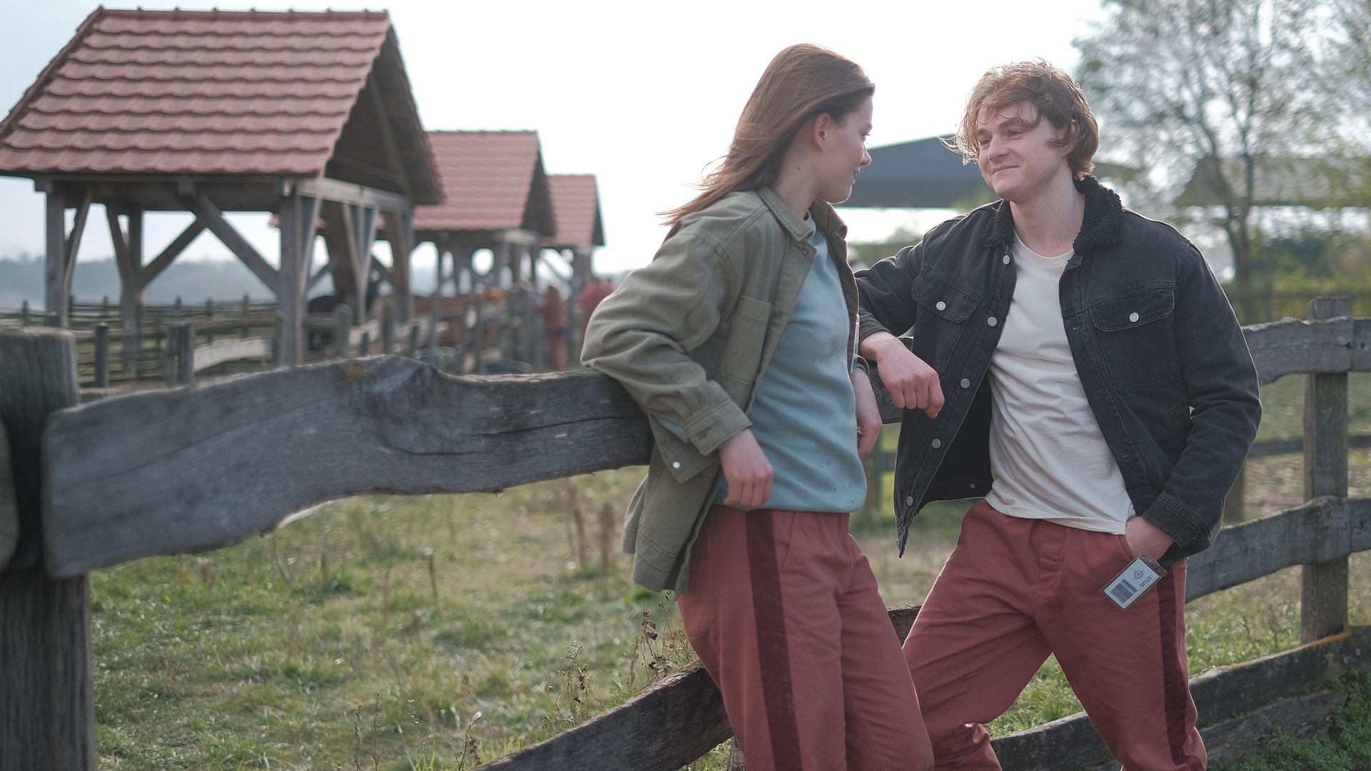 "Fallen - Vorahnung": Luce (Jessica Alexander) und Cam (Timothy Innes) lehnen entspannt an einem hölzernen Zaun und blicken sich lächelnd an. Im Hintergrund stehen mehrere Stallgebäude und Bäume.