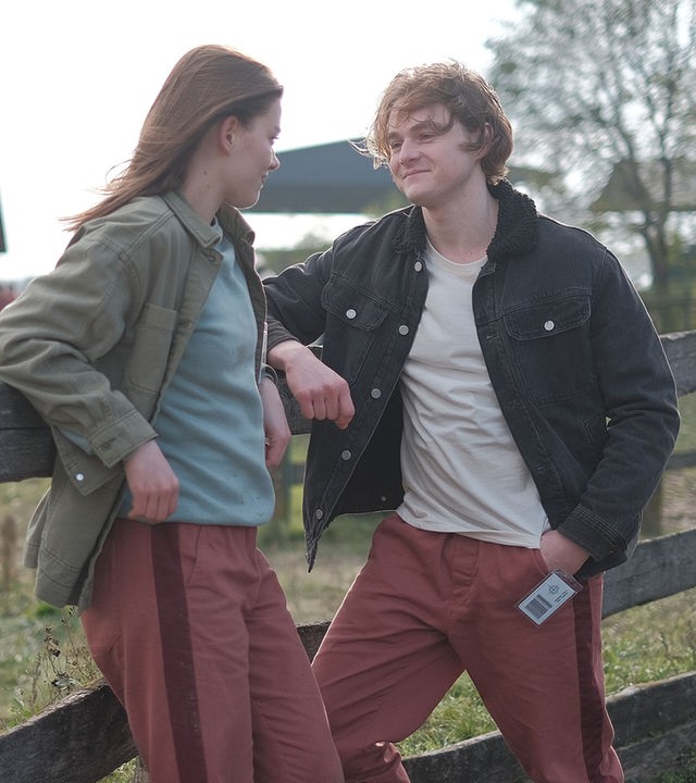 "Fallen - Vorahnung": Luce (Jessica Alexander) und Cam (Timothy Innes) lehnen entspannt an einem hölzernen Zaun und blicken sich lächelnd an. Im Hintergrund stehen mehrere Stallgebäude und Bäume.