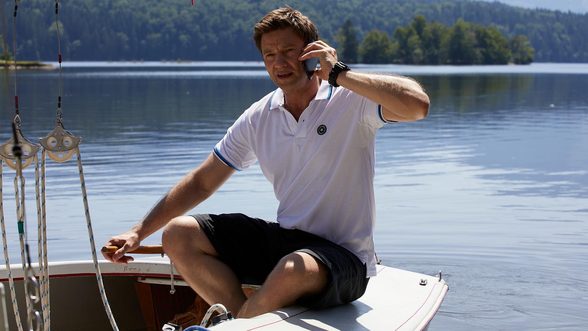 "Die Rosenheim-Cops - Ein Mord mit Vorermittlungen": Kommissar Hansen (Igor Jeftic) sitzt in einem Segelboot auf einem See und telefoniert. Im Hintergrund sind Berge und Wälder zu sehen.