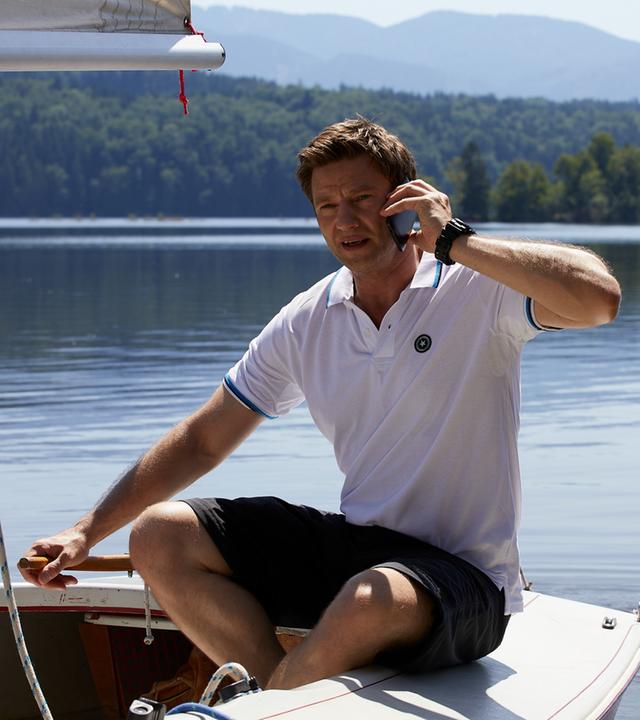 "Die Rosenheim-Cops - Ein Mord mit Vorermittlungen": Kommissar Hansen (Igor Jeftic) sitzt in einem Segelboot auf einem See und telefoniert. Im Hintergrund sind Berge und Wälder zu sehen.