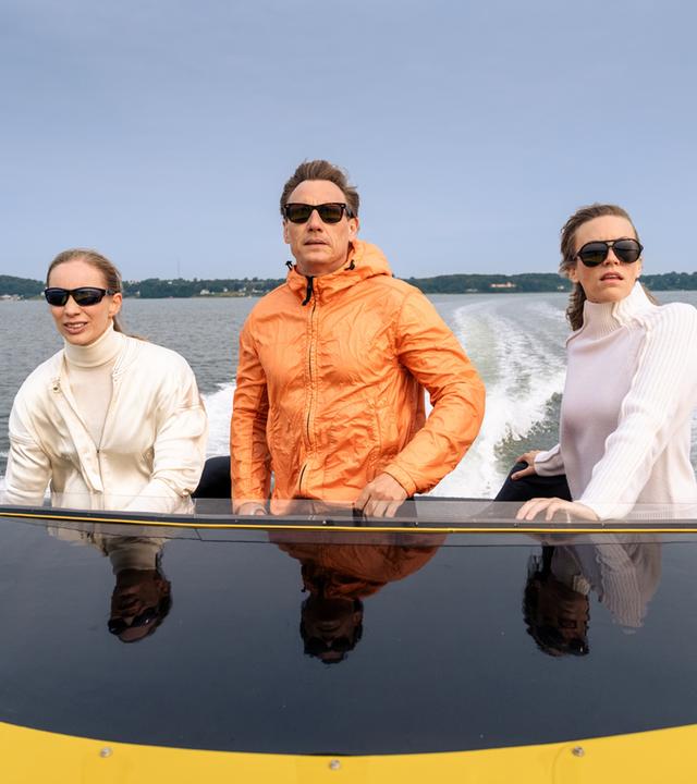 Neiss (Patrick Kalupa) trägt eine orangefarbene Bomberjacke und Sonnenbrille. Er steht zwischen zwei Frauen, Bootsführerin (Cosma Dujat) und Passagierin (Ilka Selmer), auf einem gelben Speedboot und rast über das Wasser. Beide Frauen tragen weiße Kleidung und dunkle Sonnenbrillen.