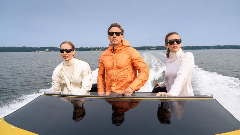 Neiss (Patrick Kalupa) trägt eine orangefarbene Bomberjacke und Sonnenbrille. Er steht zwischen zwei Frauen, Bootsführerin (Cosma Dujat) und Passagierin (Ilka Selmer), auf einem gelben Speedboot und rast über das Wasser. Beide Frauen tragen weiße Kleidung und dunkle Sonnenbrillen.