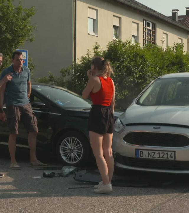 "Vorsicht, Falle! - Autobumser": Eine junge Autofahrerin steht vor ihrem Wagen, der von der Seite von einem anderen Wagen angefahren wurde. Zwei Männer reden auf sie ein. 