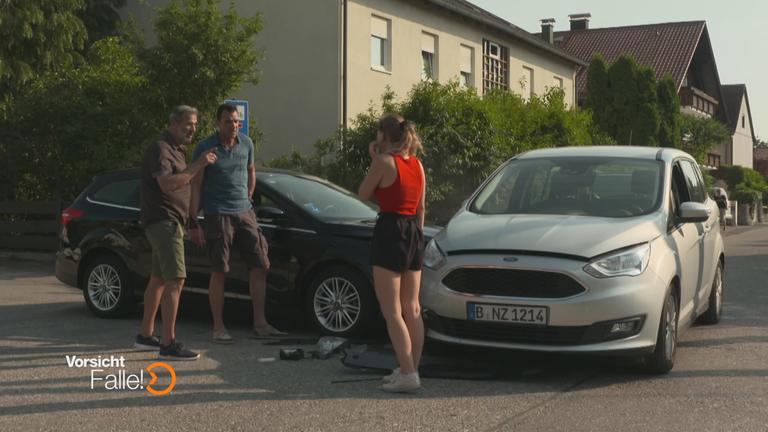"Vorsicht, Falle! - Autobumser": Eine junge Autofahrerin steht vor ihrem Wagen, der von der Seite von einem anderen Wagen angefahren wurde. Zwei Männer reden auf sie ein. 