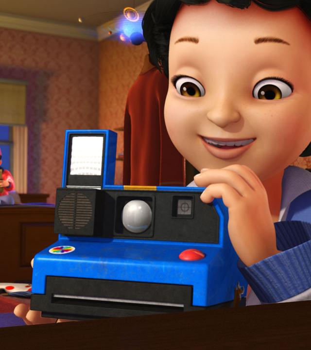 Michael mit einer blauen Sofortbild-Kamera. John sitzt im Hintergrund auf dem Bett und liest in einem Buch.