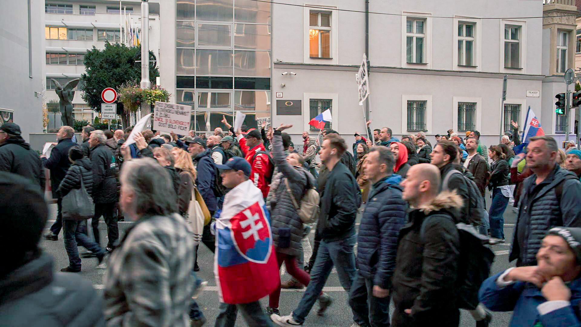 Das Bild zeigt eine Menschenmenge, die auf einer Straße demonstriert. Die Teilnehmerinnen und Teilnehmer tragen überwiegend dunkle winterliche Kleidung und einige von ihnen halten Schilder in den Händen. Es sind auch Fahnen sichtbar, darunter eine mit dem slowakischen Wappen.   Im Hintergrund sind moderne Gebäude zu sehen, darunter Glasfassaden, die das Licht reflektieren. Es wirkt, als wäre die Stimmung in der Menschenmenge aktiv und engagiert, da viele Personen ihre Arme heben und mit Schildern gestikulieren.   Die Szene spielt sich in einer städtischen Umgebung ab, wahrscheinlich in einer Hauptstadt, und deutet auf eine politische Demonstration hin, die sich gegen die Regierung richtet. Die Architektur der Gebäude ist schlicht, mit strukturierten Wänden und einer Mischung aus modernen und traditionellen Elementen.