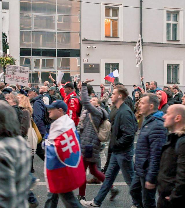 Das Bild zeigt eine Menschenmenge, die auf einer Straße demonstriert. Die Teilnehmerinnen und Teilnehmer tragen überwiegend dunkle winterliche Kleidung und einige von ihnen halten Schilder in den Händen. Es sind auch Fahnen sichtbar, darunter eine mit dem slowakischen Wappen.   Im Hintergrund sind moderne Gebäude zu sehen, darunter Glasfassaden, die das Licht reflektieren. Es wirkt, als wäre die Stimmung in der Menschenmenge aktiv und engagiert, da viele Personen ihre Arme heben und mit Schildern gestikulieren.   Die Szene spielt sich in einer städtischen Umgebung ab, wahrscheinlich in einer Hauptstadt, und deutet auf eine politische Demonstration hin, die sich gegen die Regierung richtet. Die Architektur der Gebäude ist schlicht, mit strukturierten Wänden und einer Mischung aus modernen und traditionellen Elementen.