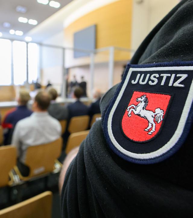 Ausschnitt aus dem Gerichtssaal. Es sind einige Besucher anwesend. Außerdem ist Personal der Justiz zu sehen.