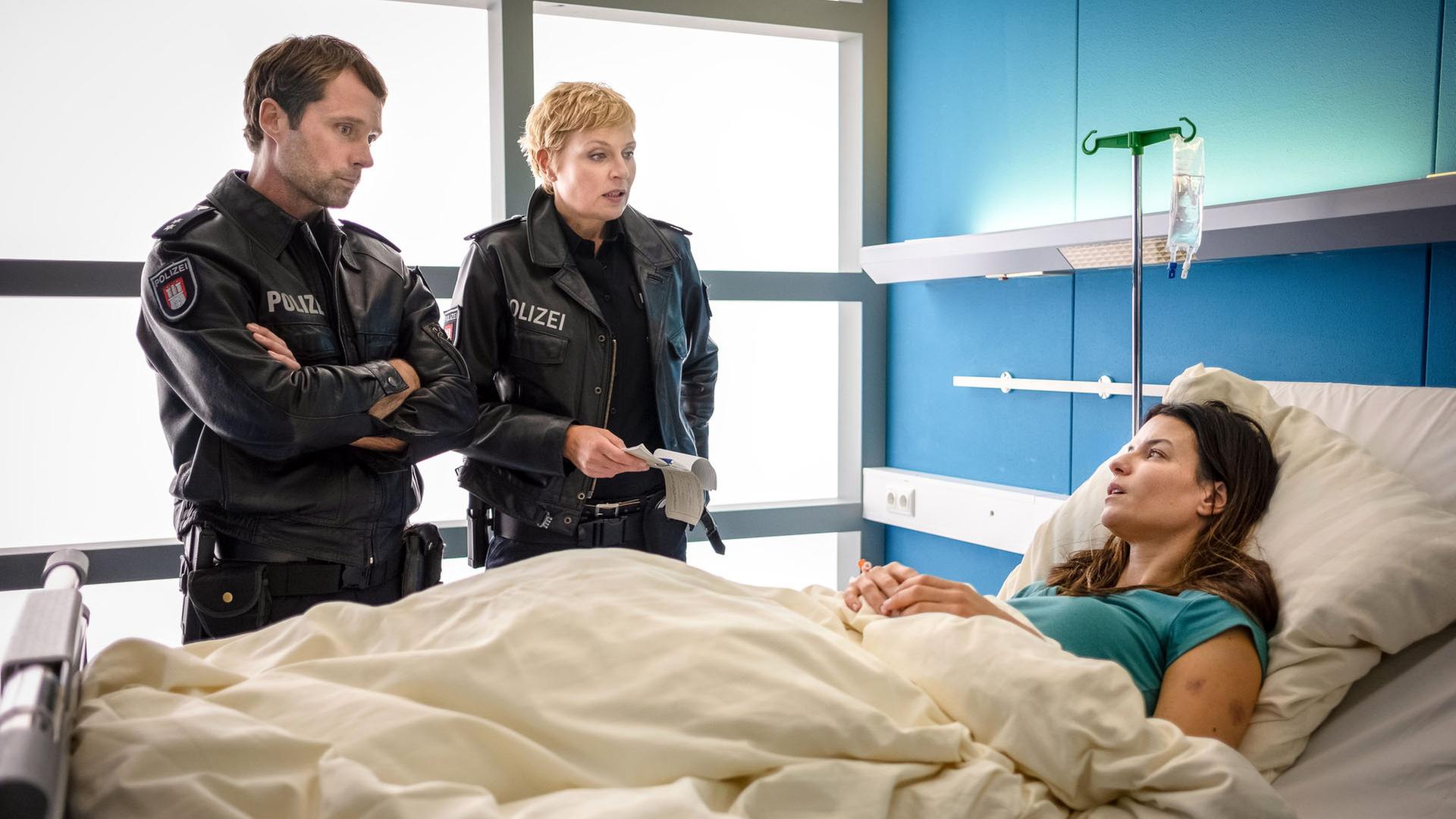 "Notruf Hafenkante - Wach auf!": Die Polizeibeamten Mattes Seeler (Matthias Schloo) und Melanie Hansen (Sanna Englund) stehen nebeneinander an der rechten Seite eines Krankenbettes, in dem Sofie Blum (Anna Lena Class) liegt. Die Beamten sehen Sofie an.