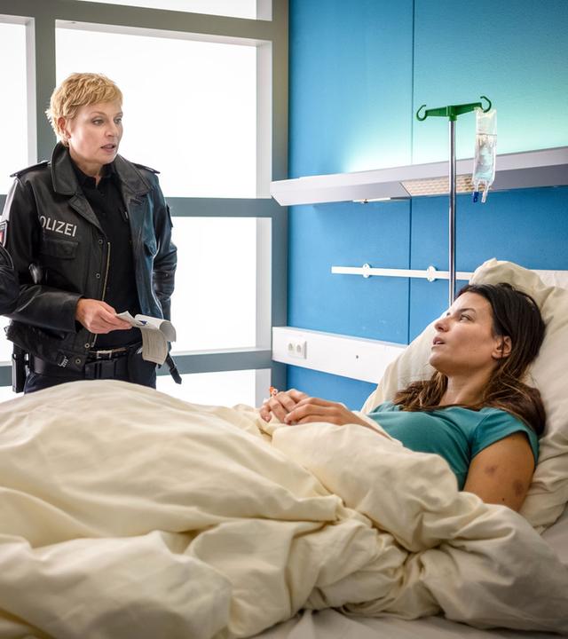 "Notruf Hafenkante - Wach auf!": Die Polizeibeamten Mattes Seeler (Matthias Schloo) und Melanie Hansen (Sanna Englund) stehen nebeneinander an der rechten Seite eines Krankenbettes, in dem Sofie Blum (Anna Lena Class) liegt. Die Beamten sehen Sofie an.