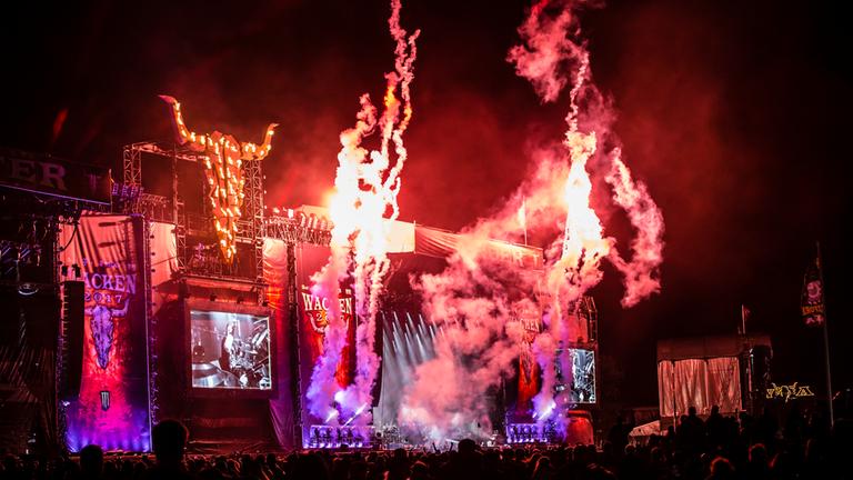Die Bühne des Wacken Open Air ist aus Publikumsperspektive zu sehen. Sie ist in rötliches und lilafarbenes Licht getaucht. Mehrere Nebelschwaden steigen vor der Bühne in die Luft.