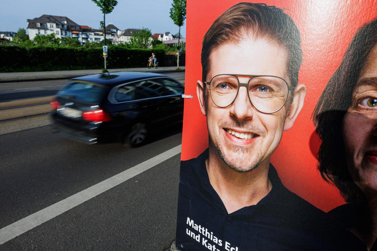 Ein rotes Wahlplakat von SPD-Kandidat Matthias Ecke, einem Mann mit Dreitagebart und großer Brille, ist am Straßenrand zu sehen.