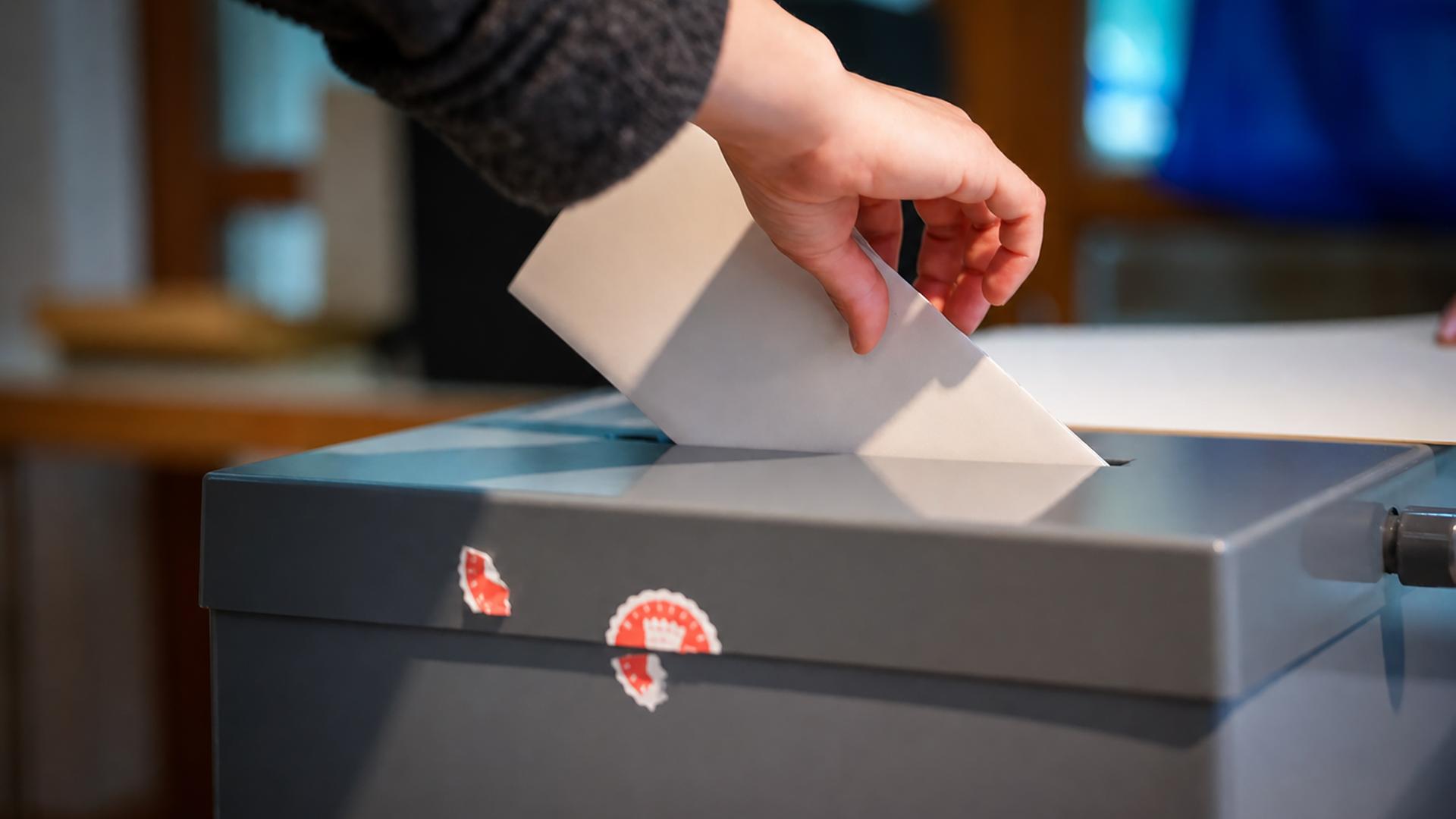 Eine Hand schiebt ein gefaltetes Papier in den Schlitz einer Wahlurne