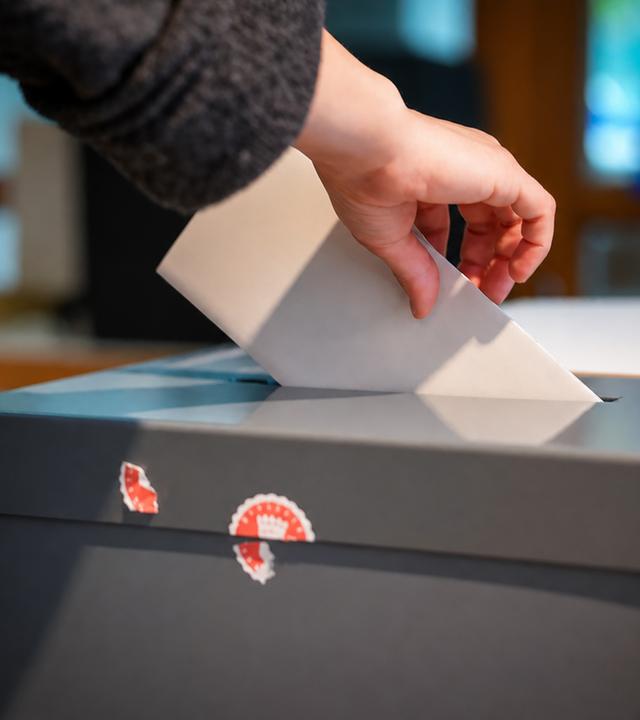 Eine Hand schiebt ein gefaltetes Papier in den Schlitz einer Wahlurne