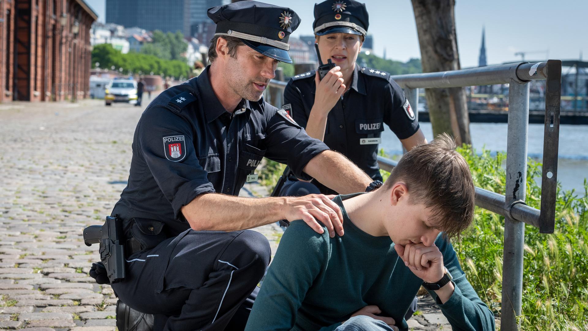Malte Hinrichs (Leon Blaschke) sitzt auf dem Kopfsteinpflaster neben einem Geländer. Der Polizist Mattes Seeler (Matthias Schloo) sitzt in der Hocke hinter ihm und hält ihn am Oberkörper. Hinter Mattes sitzt seine Kollegin Melanie Hansen (Sanna Englund) in der Hocke. Sie spricht in ihr Funkgerät.
