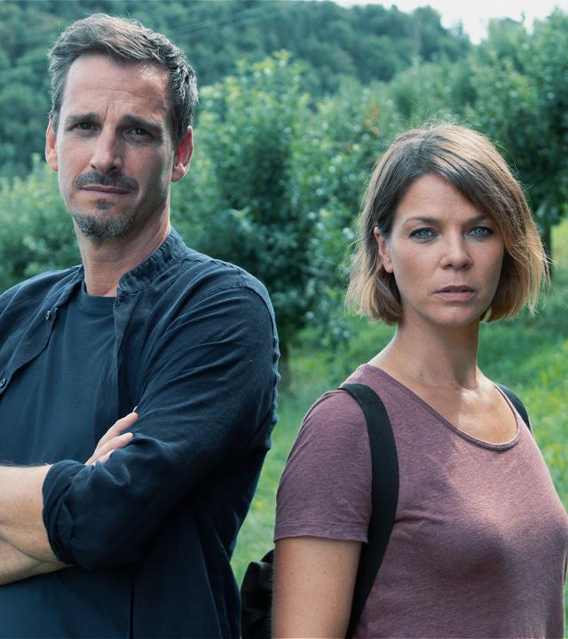Die Kommissare Konrad Diener (Max von Thun) und Maris Bächle (Jessica Schwarz) stehen mit den Rücken zueinander auf einer Wiese und schauen in die Kamera. Hinter ihnen erstreckt sich eine Waldlandschaft mit dichten Bäumen.