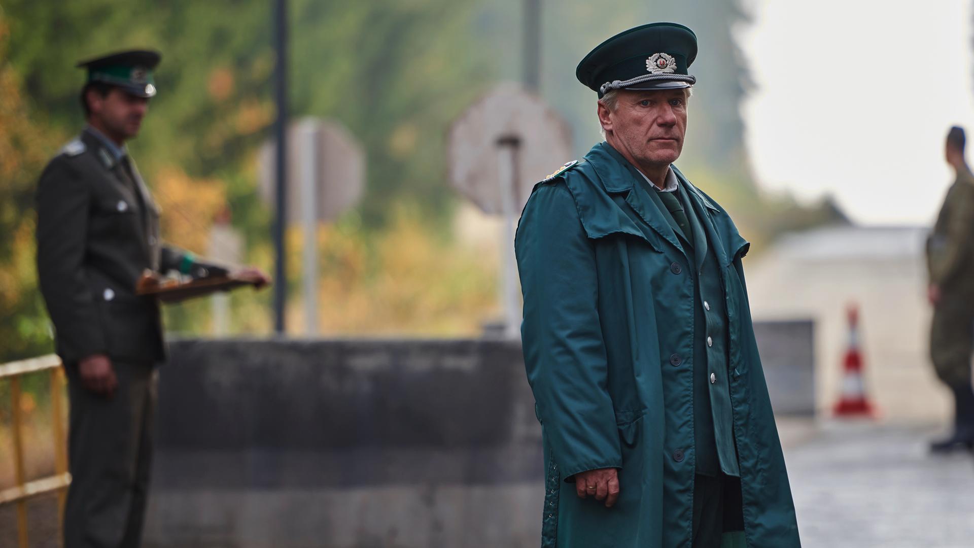 Lothar Wieditz (Jörg Schüttauf) steht in grüner Polizeiuniform auf einer Straße, die von bewaffneten Männern bewacht wird.