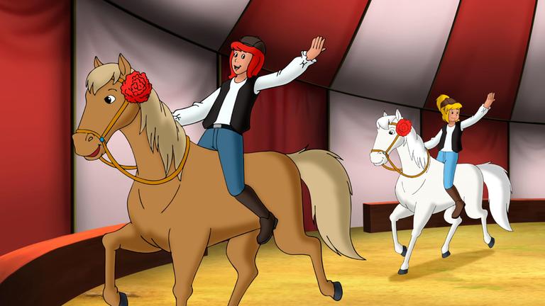 Tina (vorne links) und Bibi (hinten rechts) reiten auf Pferden in einer Zirkusmanege. Beide halten sich mit einer Hand am Pferd fest, die andere haben sie weit in die Luft gestreckt und lachen dabei.