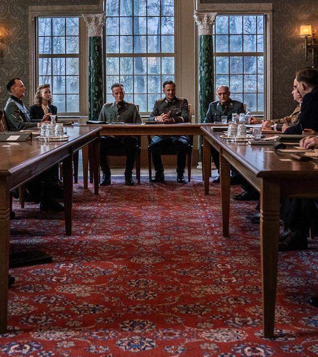 "Die Wannseekonferenz": Im repräsentativen Besprechungssaal sitzen die Teilnehmer an einem Tisch in U-Form. Auf dem Tisch liegen Besprechungsunterlagen. Auf Tabletts stehen kristallene Gläser und Wasserkaraffen. Die Teilnehmer wenden ihre Blicke zu Reinhard Heydrich (Philipp Hochmair), der an der Stirnseite des Tisches sitzt und die Besprechung eröffnet hat.