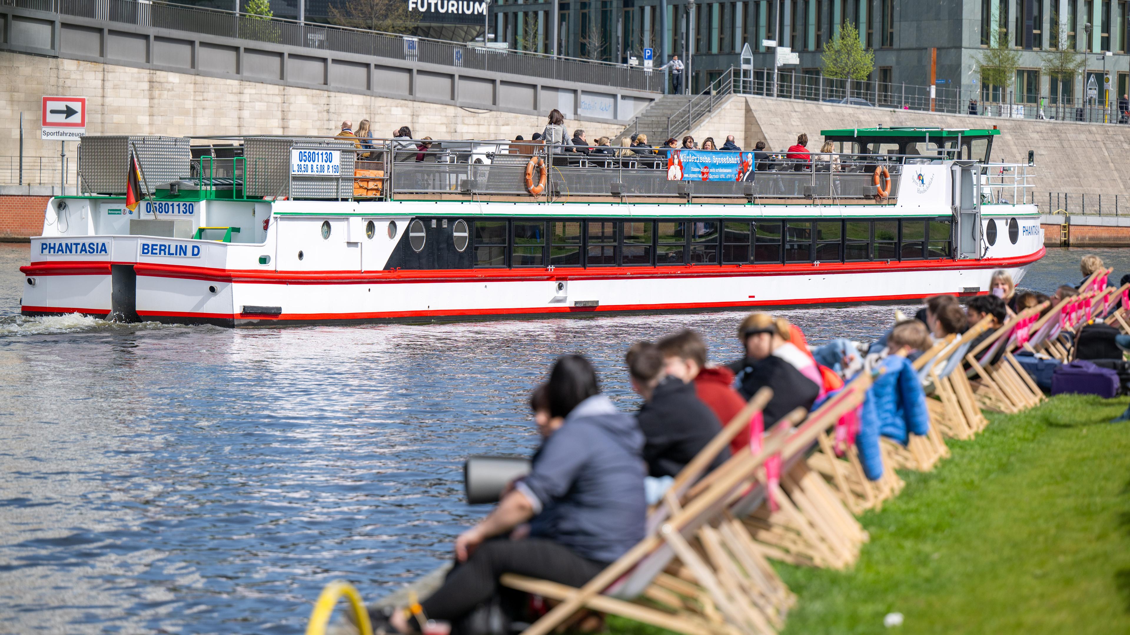 Berlin: Menschen sitzen beim sonnigen Wetter am Spreeufer, während im Hintergrund ein Ausflugsschiff vorbeifährt.
