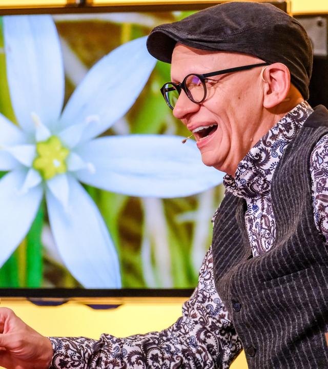 Das Bild zeigt einen lächelnden Mann, der in einem Studio sitzt. Er trägt eine schwarze Mütze und eine Brille mit runden Gläsern. Seine Kleidung besteht aus einem gemusterten Hemd mit floralem Design und einer dunklen Weste. Vor ihm steht ein Glas Wasser. In der Hand hält er einen Stift. Im Hintergrund ist ein großer Bildschirm zu sehen, auf dem eine Nahaufnahme einer weißen Blume zu sehen ist, die auf einem grünen Hintergrund abgebildet ist. Die Umgebung wirkt hell und freundlich, und der Mann sieht aus, als würde er etwas unterhaltsames oder interessantes erzählen.