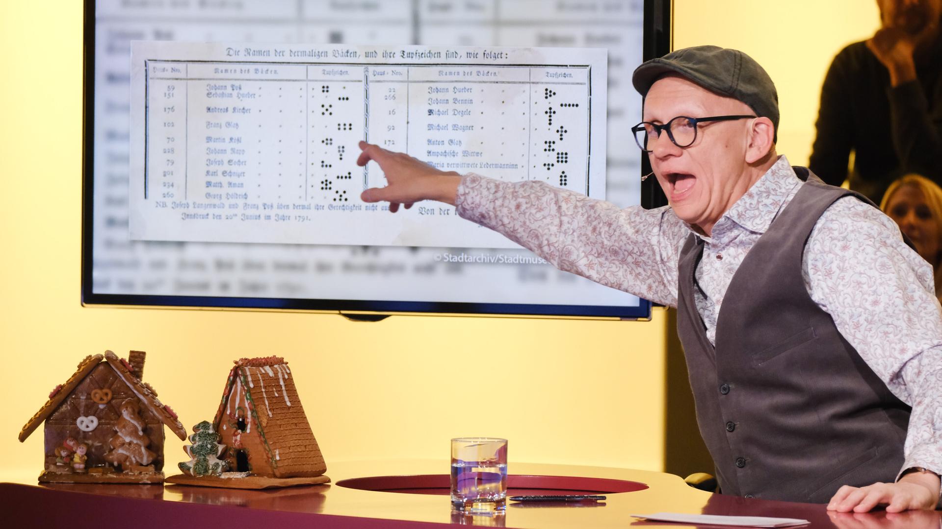 Das Bild zeigt eine Szene aus einer Fernsehshow. Im Vordergrund steht ein runder Tisch mit zwei kleinen Lebkuchenhäusern, die dekoriert sind. Eines ist halbrund und das andere hat eine spitze Dachform. Auf dem Tisch steht zudem ein Glas Wasser und ein Stift.  Hinter dem Tisch ist ein großer Bildschirm zu sehen, auf dem ein Dokument dargestellt ist, das anscheinend Daten oder Namen auflistet.   Ein Mann, der aufgeregt wirkt, steht an dem Tisch und gestikuliert mit einer Hand, während er auf den Bildschirm zeigt. Er trägt ein Hemd mit einem blumigen Muster, eine graue Weste und eine Mütze. Im Hintergrund sind mehrere Zuschauer unscharf zu erkennen, die die Szene beobachten. Die Beleuchtung ist warm und einladend.