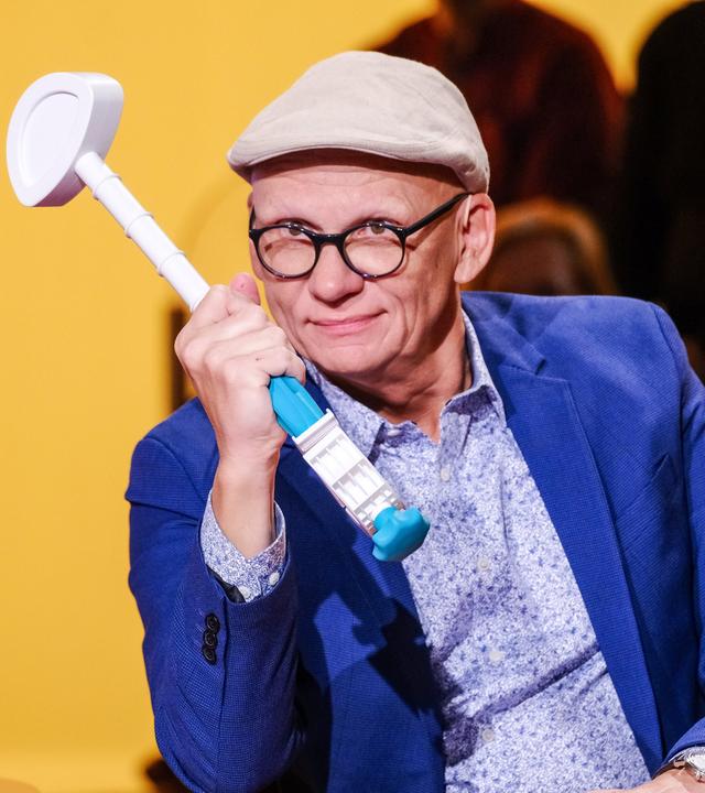 Auf dem Bild ist ein Mann zu sehen, der einen hellen Hut trägt und eine Brille mit schwarzem Gestell hat. Er sitzt an einem Tisch und hält einen Gegenstand in der Hand, der aussieht wie ein Spielzeug oder ein Mikrofon mit einer blauen und weißen Farbe. Der Mann trägt ein blaues Sakko über einem hellen, gemusterten Hemd. Im Hintergrund ist eine wandfarbene gelb-orange Farbe zu erkennen, sowie einige undeutliche Silhouetten von weiteren Personen. Der Gesamtkomplex vermittelt eine fröhliche und lockere Atmosphäre.