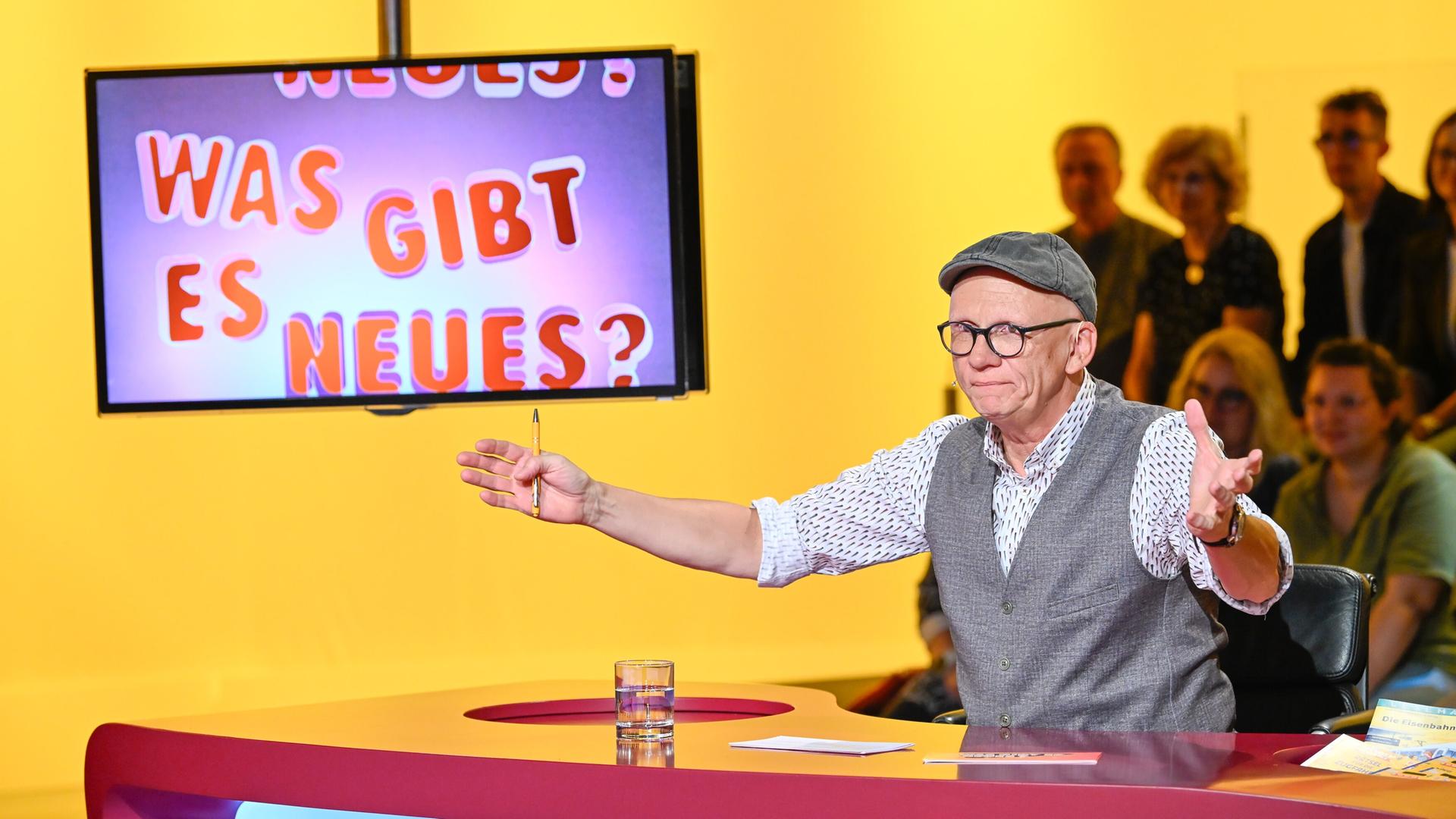 Das Bild zeigt eine Fernsehsendung mit dem Titel „Was gibt es Neues?“. Im Vordergrund steht ein Moderator, der Oliver Baier ist. Er trägt eine graue Weste über einem weißen Hemd mit buntem Muster und eine graue Schirmmütze. Seine Brille hat dünne, schwarze Rahmen. Er gestikuliert mit einer Hand, während er auf einem Tisch sitzt, der eine rote Oberfläche hat. Auf dem Tisch steht ein Glas mit Wasser und einige Unterlagen.  Im Hintergrund ist ein Bildschirm sichtbar, der den Titel der Show in bunten Buchstaben anzeigt: „NEUES? WAS GIBT ES NEUES?“. Der Hintergrund des Bildes ist gelb. Hinter dem Moderator sind mehrere Zuschauer zu sehen, die teilweise unscharf sind. Sie scheinen interessiert zuzuschauen.