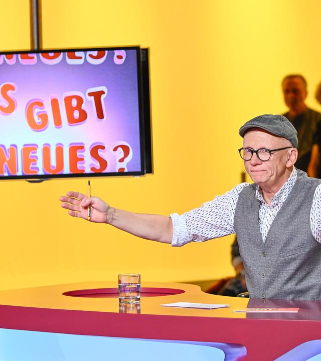 Das Bild zeigt eine Fernsehsendung mit dem Titel „Was gibt es Neues?“. Im Vordergrund steht ein Moderator, der Oliver Baier ist. Er trägt eine graue Weste über einem weißen Hemd mit buntem Muster und eine graue Schirmmütze. Seine Brille hat dünne, schwarze Rahmen. Er gestikuliert mit einer Hand, während er auf einem Tisch sitzt, der eine rote Oberfläche hat. Auf dem Tisch steht ein Glas mit Wasser und einige Unterlagen.  Im Hintergrund ist ein Bildschirm sichtbar, der den Titel der Show in bunten Buchstaben anzeigt: „NEUES? WAS GIBT ES NEUES?“. Der Hintergrund des Bildes ist gelb. Hinter dem Moderator sind mehrere Zuschauer zu sehen, die teilweise unscharf sind. Sie scheinen interessiert zuzuschauen.