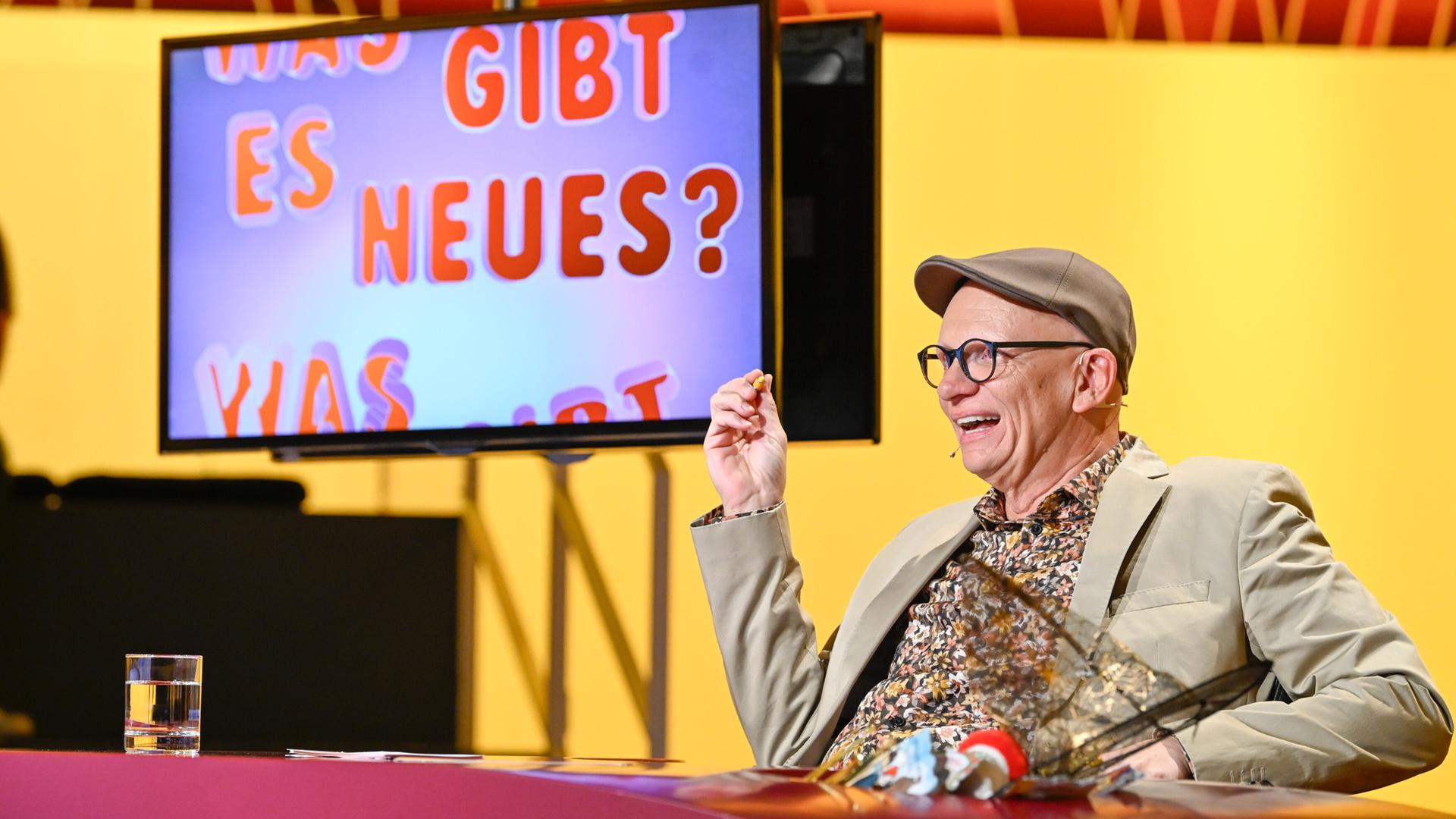 Das Bild zeigt eine Fernsehszene aus der Show "Was gibt es Neues?". Im Vordergrund sitzt ein Mann, der fröhlich aussieht und in einem beigen Anzug mit einem bunten, gemusterten Hemd und einer grauen Mütze gekleidet ist. Er hält in einer Hand einen kleinen Snack und gestikuliert, während er lacht oder spricht.   Hinter ihm ist ein großer Bildschirm zu sehen, auf dem in bunten Buchstaben der Text „WAS GIBT ES NEUES?“ zu lesen ist. Der Hintergrund der Szene ist in warmen Gelbtönen gehalten. Auf dem Tisch vor dem Mann steht ein Glas mit klarem Wasser. Die gesamte Atmosphäre wirkt freundlich und unterhaltsam.
