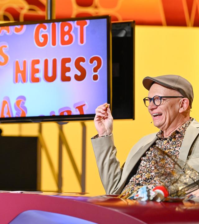 Das Bild zeigt eine Fernsehszene aus der Show "Was gibt es Neues?". Im Vordergrund sitzt ein Mann, der fröhlich aussieht und in einem beigen Anzug mit einem bunten, gemusterten Hemd und einer grauen Mütze gekleidet ist. Er hält in einer Hand einen kleinen Snack und gestikuliert, während er lacht oder spricht.   Hinter ihm ist ein großer Bildschirm zu sehen, auf dem in bunten Buchstaben der Text „WAS GIBT ES NEUES?“ zu lesen ist. Der Hintergrund der Szene ist in warmen Gelbtönen gehalten. Auf dem Tisch vor dem Mann steht ein Glas mit klarem Wasser. Die gesamte Atmosphäre wirkt freundlich und unterhaltsam.