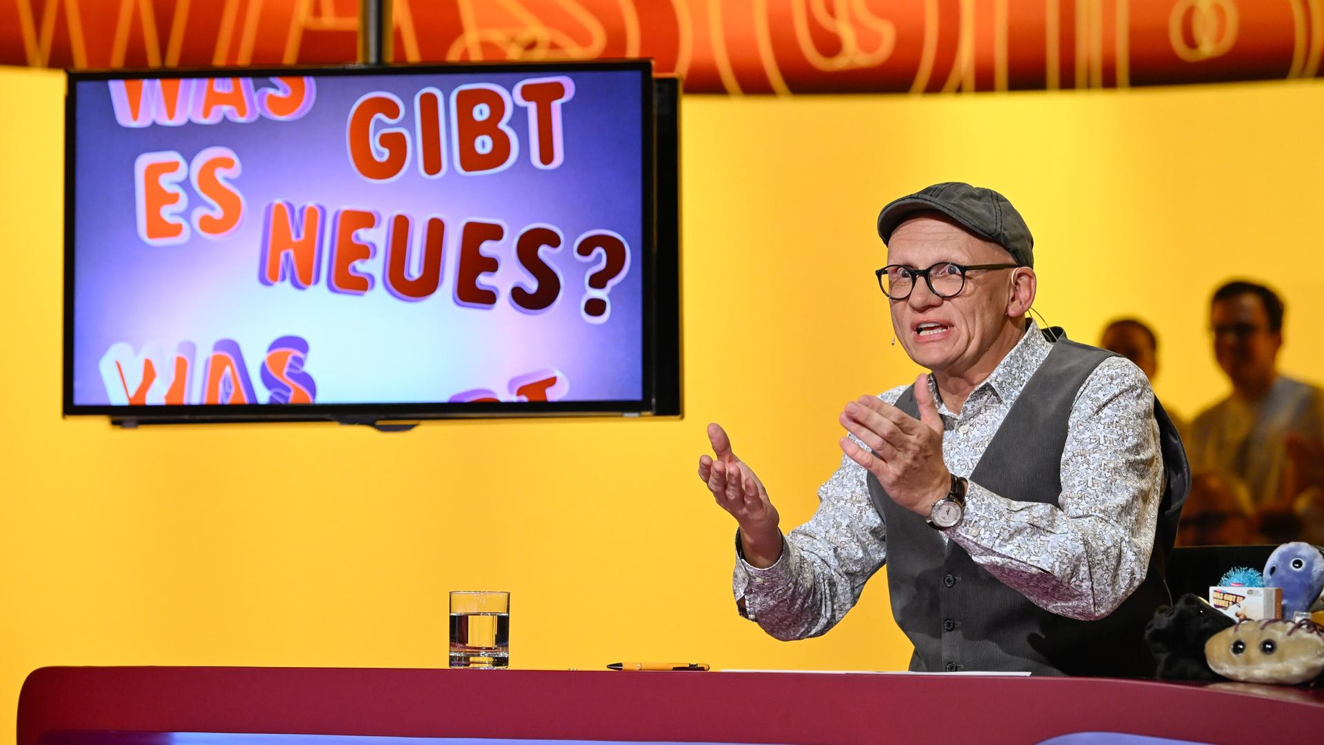 Das Bild zeigt eine Studio-Szene aus der Fernsehsendung "Was gibt es Neues?". Im Vordergrund sitzt ein Mann, der Oliver Baier ist. Er trägt eine graue Mütze, eine brille und ein gemustertes Hemd unter einer grauen Weste. Seine Hände sind erhoben, als ob er einen Punkt erklärt. Vor ihm steht ein Tisch, auf dem ein Glas Wasser und ein Schreibgerät liegen. Rechts auf dem Tisch sind einige farbige Plüschfiguren zu sehen.  Im Hintergrund ist ein Bildschirm, auf dem der Text "Was gibt es Neues?" in bunten Buchstaben zu sehen ist. Die Wand hinter Oliver Baier hat einen gelben Farbton mit einem abstrakten Muster. Im Hintergrund sind einige schemenhafte Personen zu erkennen, die im Publikum sitzen.   Die Gesamtatmosphäre vermittelt eine lebhafte und unterhaltsame Stimmung, die typisch für eine Quiz- oder Unterhaltungssendung ist.