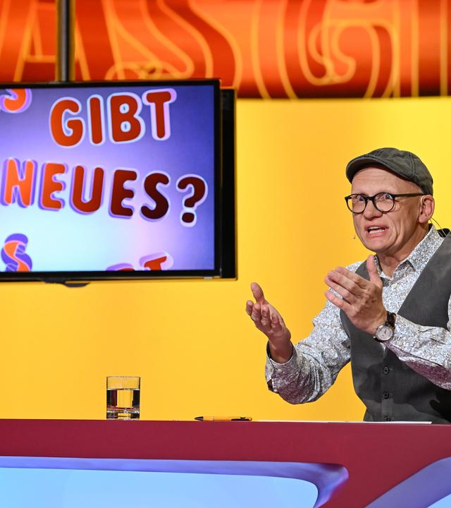 Das Bild zeigt eine Studio-Szene aus der Fernsehsendung "Was gibt es Neues?". Im Vordergrund sitzt ein Mann, der Oliver Baier ist. Er trägt eine graue Mütze, eine brille und ein gemustertes Hemd unter einer grauen Weste. Seine Hände sind erhoben, als ob er einen Punkt erklärt. Vor ihm steht ein Tisch, auf dem ein Glas Wasser und ein Schreibgerät liegen. Rechts auf dem Tisch sind einige farbige Plüschfiguren zu sehen.  Im Hintergrund ist ein Bildschirm, auf dem der Text "Was gibt es Neues?" in bunten Buchstaben zu sehen ist. Die Wand hinter Oliver Baier hat einen gelben Farbton mit einem abstrakten Muster. Im Hintergrund sind einige schemenhafte Personen zu erkennen, die im Publikum sitzen.   Die Gesamtatmosphäre vermittelt eine lebhafte und unterhaltsame Stimmung, die typisch für eine Quiz- oder Unterhaltungssendung ist.