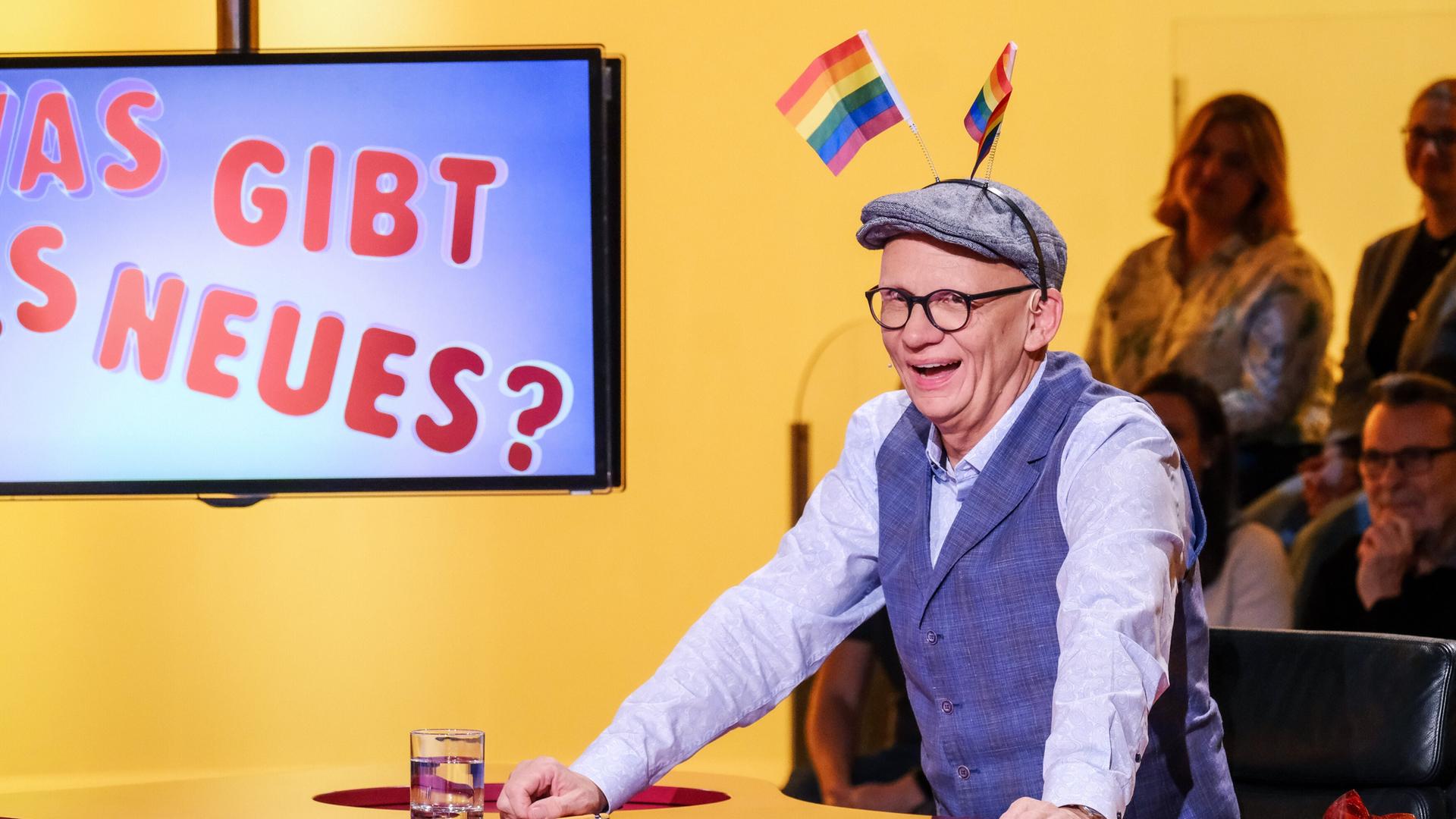Das Bild zeigt einen Mann, der an einem Tisch sitzt und lächelt. Er trägt eine graue Mütze und eine Weste über einem hellen Hemd. Auf seiner Mütze sind zwei kleine Regenbogenflaggen befestigt, die hochstehen. Vor ihm steht ein Glas mit Wasser und auf dem Tisch liegt ein Schreibblock. Im Hintergrund ist eine großformatige Bildschirmanzeige, auf der in bunten Buchstaben die Frage "Was gibt es Neues?" zu lesen ist. Die Umgebung hat eine gelbe Wandfarbe, und im Hintergrund sind mehrere Personen zu sehen, die anscheinend der Veranstaltung folgen.