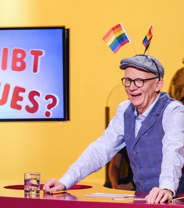 Das Bild zeigt einen Mann, der an einem Tisch sitzt und lächelt. Er trägt eine graue Mütze und eine Weste über einem hellen Hemd. Auf seiner Mütze sind zwei kleine Regenbogenflaggen befestigt, die hochstehen. Vor ihm steht ein Glas mit Wasser und auf dem Tisch liegt ein Schreibblock. Im Hintergrund ist eine großformatige Bildschirmanzeige, auf der in bunten Buchstaben die Frage "Was gibt es Neues?" zu lesen ist. Die Umgebung hat eine gelbe Wandfarbe, und im Hintergrund sind mehrere Personen zu sehen, die anscheinend der Veranstaltung folgen.