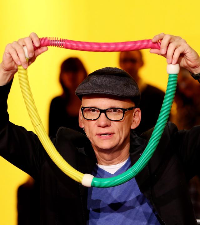 Ein Mann hält sich eine bunte, breite Kette vor den Kopf.