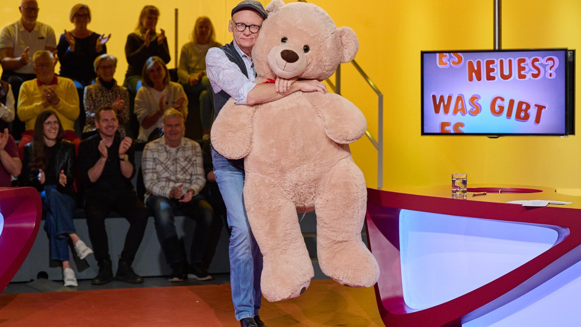 Auf dem Bild ist ein Moderator zu sehen, der einen großen, plüschigen Teddy in den Armen hält. Der Moderator trägt ein Hemd mit einem bunten Muster, eine Weste und eine Mütze. Im Hintergrund sitzt ein Publikum auf gestuften Plätzen und applaudiert.  Das Setting ist eine Fernsehsendung mit einem farbenfrohen, modern gestalteten Set. An einer Wand hinter dem Moderator ist ein Bildschirm mit dem Schriftzug „ES NEUES? WAS GIBT ES“ zu sehen. Der Tisch, an dem der Moderator steht, hat eine kurvenreiche, rote Oberfläche und ist teilweise transparent blau beleuchtet. Auf dem Tisch liegen einige Notizen und ein Glas.   Die gesamte Szenerie strahlt eine fröhliche und unterhaltsame Atmosphäre aus.