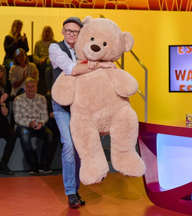 Auf dem Bild ist ein Moderator zu sehen, der einen großen, plüschigen Teddy in den Armen hält. Der Moderator trägt ein Hemd mit einem bunten Muster, eine Weste und eine Mütze. Im Hintergrund sitzt ein Publikum auf gestuften Plätzen und applaudiert.  Das Setting ist eine Fernsehsendung mit einem farbenfrohen, modern gestalteten Set. An einer Wand hinter dem Moderator ist ein Bildschirm mit dem Schriftzug „ES NEUES? WAS GIBT ES“ zu sehen. Der Tisch, an dem der Moderator steht, hat eine kurvenreiche, rote Oberfläche und ist teilweise transparent blau beleuchtet. Auf dem Tisch liegen einige Notizen und ein Glas.   Die gesamte Szenerie strahlt eine fröhliche und unterhaltsame Atmosphäre aus.