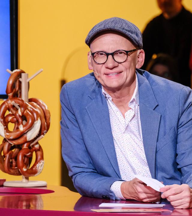 Das Bild zeigt einen Mann, der am Tisch vor einem orangefarbenen Hintergrund sitzt. Er trägt eine graue Mütze und eine Brille mit dicken, schwarzen Rahmen. Seine Kleidung besteht aus einem hellblauen Blazer über einem weißen Hemd mit bunten Punkten. Auf dem Tisch vor ihm liegen einige Zettel und ein Glas mit Wasser.  Neben ihm steht eine Art Ständer mit mehreren Brezeln, die übereinander gestapelt sind. Im Hintergrund sind mehrere Personen zu sehen, die an einer Art Publikumssitzplatz sitzen. Der Gesamtaufbau der Szene scheint zu einem Fernsehformat zu gehören, das sich mit dem Thema „Brezel“ beschäftigt.