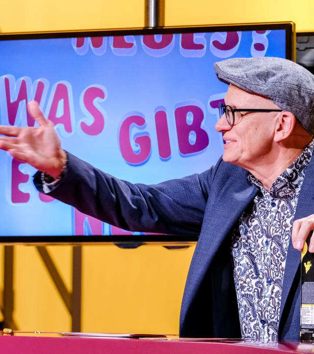 Das Bild zeigt einen Mann, der im Sitzen mit einer Hand gestikuliert. Er trägt eine graue Mütze und eine Brille. Sein Oberteil ist ein Musterhemd in verschiedenen Farben, über dem er einen dunkelblauen Blazer trägt. Vor ihm steht ein Tisch mit verschiedenen Objekten, darunter ein Glas Wasser und eine Packung mit Snacks. Im Hintergrund ist ein Bildschirm zu sehen, auf dem in bunten Buchstaben der Text "Was gibt es Neues?" steht. Der Raum hat eine gelbe Wandfarbe, und es sind einige strukturelle Elemente sichtbar, die die Umgebung gestalten.