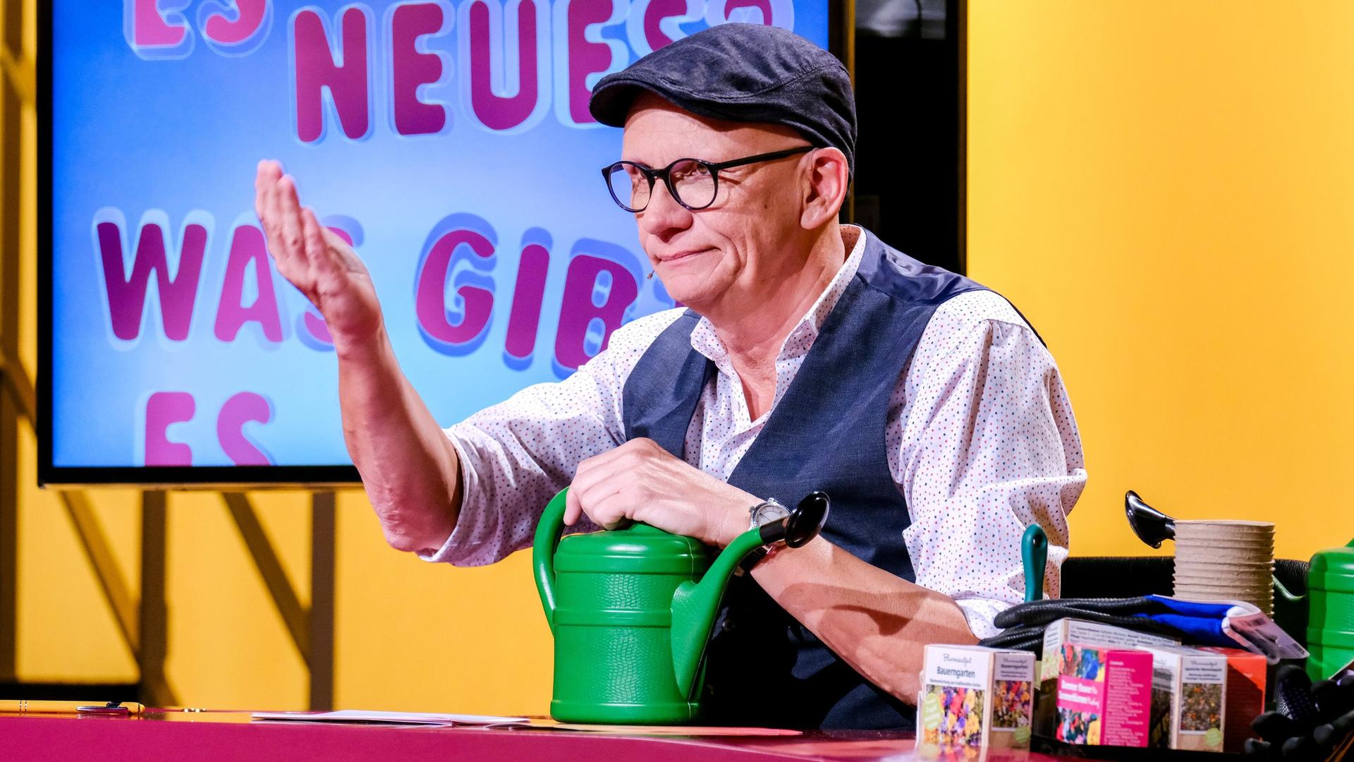Das Bild zeigt einen Mann, der an einem Tisch sitzt und eine grüne Gießkanne in der Hand hält. Er trägt eine graue Mütze, runde Brille und ein Hemd mit weißen Punkten. Seine Weste ist dunkelblau. Der Hintergrund ist in Gelbtönen gehalten und zeigt einen Bildschirm mit der Aufschrift „ES NEUES WAS GIBT“. Auf dem Tisch sind verschiedene Gegenstände zu sehen, darunter eine weitere Gießkanne, eine kleinere Kiste mit bunter Verpackung und einige andere Utensilien. Der Mann hat eine Hand erhoben, wahrscheinlich in einem Gestus des Erklärens oder des Gesprächs.