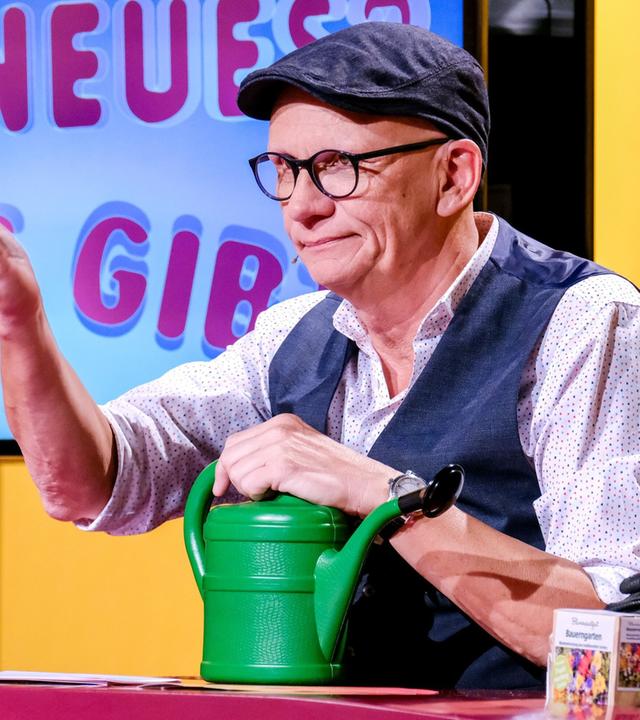 Das Bild zeigt einen Mann, der an einem Tisch sitzt und eine grüne Gießkanne in der Hand hält. Er trägt eine graue Mütze, runde Brille und ein Hemd mit weißen Punkten. Seine Weste ist dunkelblau. Der Hintergrund ist in Gelbtönen gehalten und zeigt einen Bildschirm mit der Aufschrift „ES NEUES WAS GIBT“. Auf dem Tisch sind verschiedene Gegenstände zu sehen, darunter eine weitere Gießkanne, eine kleinere Kiste mit bunter Verpackung und einige andere Utensilien. Der Mann hat eine Hand erhoben, wahrscheinlich in einem Gestus des Erklärens oder des Gesprächs.