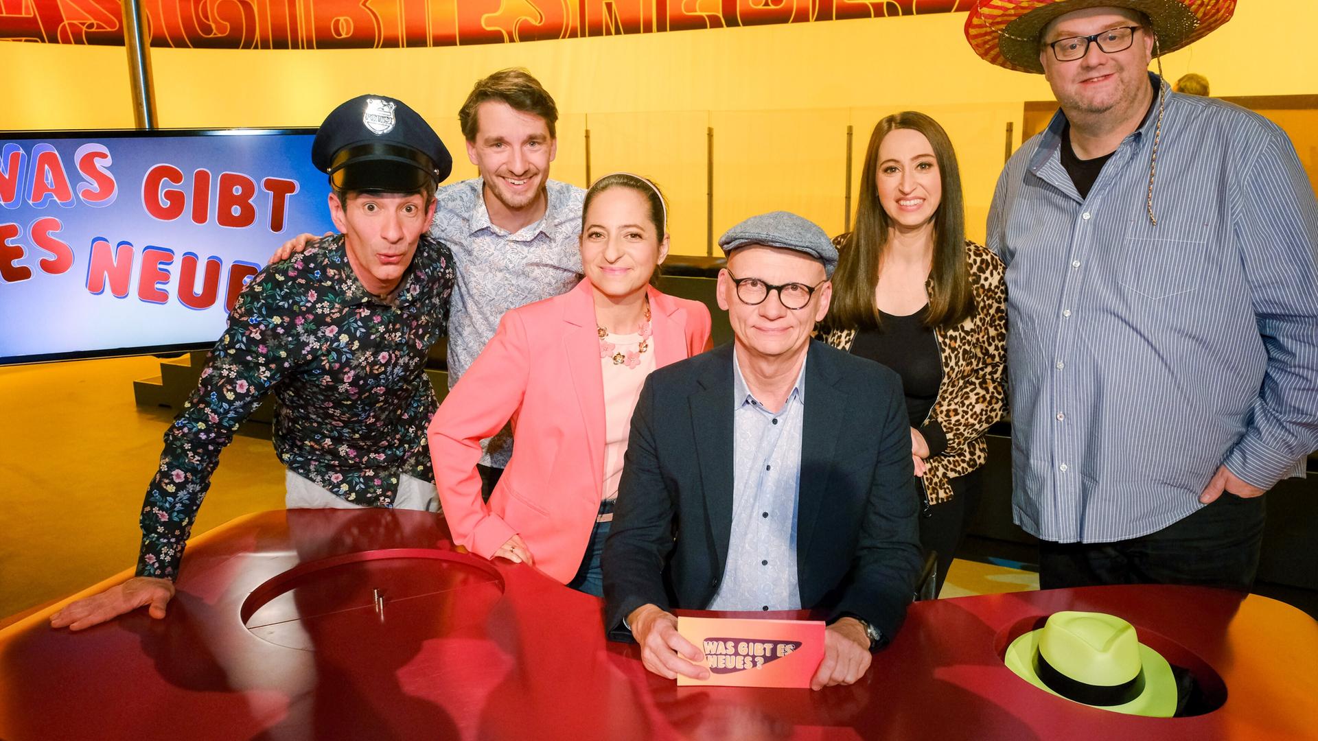 Das Bild zeigt eine Gruppe von sechs Personen, die an einem runden Tisch in einer Studioatmosphäre sitzen. Die Rückwand ist in einem leuchtenden Gelb gehalten und zeigt den Schriftzug „WAS GIBT ES NEUES?“.   Die Personen sind um den Tisch angeordnet. Links steht ein Mann in einem schwarzen Shirt mit floralen Mustern und hat eine Polizeimütze aufgesetzt. Neben ihm lächelt ein weiterer Mann in einem hellen Hemd mit Karomuster. Weiter rechts sitzt eine Frau in einem rosa Blazer, daneben ist eine Person mit einer grauen Mütze. Diese Person hält ein Kärtchen in der Hand, auf dem ebenfalls der Schriftzug „WAS GIBT ES NEUES?“ zu sehen ist. Rechts von ihm steht eine Frau in einem schwarzen Oberteil mit Leopardenmuster, gefolgt von einem Mann in einem gestreiften Hemd und einem großen Hut.  Auf dem Tisch sind zwei Hüte sichtbar, ein grüner und ein schwarzer. Die Gesamtstimmung ist freundlich und humorvoll, was durch die Gesichtsausdrücke der Abgebildeten vermittelt wird. Es scheint sich um eine Fernsehaufzeichnung zu handeln.