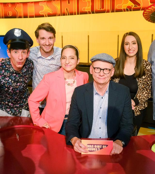 Das Bild zeigt eine Gruppe von sechs Personen, die an einem runden Tisch in einer Studioatmosphäre sitzen. Die Rückwand ist in einem leuchtenden Gelb gehalten und zeigt den Schriftzug „WAS GIBT ES NEUES?“.   Die Personen sind um den Tisch angeordnet. Links steht ein Mann in einem schwarzen Shirt mit floralen Mustern und hat eine Polizeimütze aufgesetzt. Neben ihm lächelt ein weiterer Mann in einem hellen Hemd mit Karomuster. Weiter rechts sitzt eine Frau in einem rosa Blazer, daneben ist eine Person mit einer grauen Mütze. Diese Person hält ein Kärtchen in der Hand, auf dem ebenfalls der Schriftzug „WAS GIBT ES NEUES?“ zu sehen ist. Rechts von ihm steht eine Frau in einem schwarzen Oberteil mit Leopardenmuster, gefolgt von einem Mann in einem gestreiften Hemd und einem großen Hut.  Auf dem Tisch sind zwei Hüte sichtbar, ein grüner und ein schwarzer. Die Gesamtstimmung ist freundlich und humorvoll, was durch die Gesichtsausdrücke der Abgebildeten vermittelt wird. Es scheint sich um eine Fernsehaufzeichnung zu handeln.