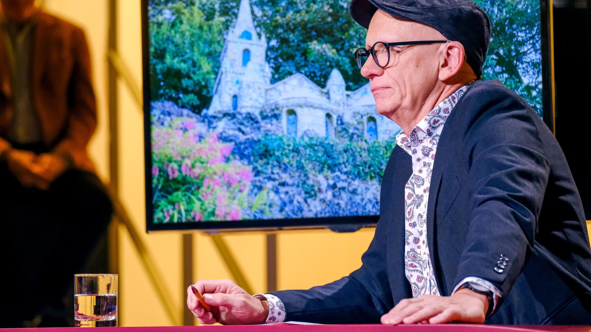 Das Bild zeigt einen Mann, der an einem Tisch sitzt und nach rechts schaut. Er trägt eine Schirmmütze und eine Brille. Sein Outfit besteht aus einem dunklen Sakko und einem bunteren Hemd mit einem Paisleymuster. Auf dem Tisch vor ihm steht ein Glas Wasser, und seine Hand hält einen Stift. Im Hintergrund ist ein Bildschirm zu sehen, der ein Bild eines architektonischen Bauwerks zeigt, das von üppigem Grün umgeben ist. Das Bauwerk hat spitze Türme und eine steinerne Fassade. Im Hintergrund ist eine weitere Person unscharf zu sehen, die anscheinend auf einem anderen Stuhl sitzt. Das gesamte Setting wirkt professionell und ist lebhaft beleuchtet.
